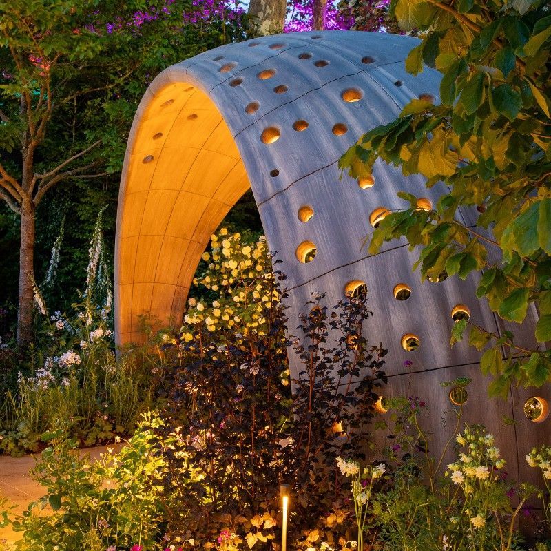 Curved gray garden sculpture with round holes, warmly lit amid lush trees and foliage at dusk