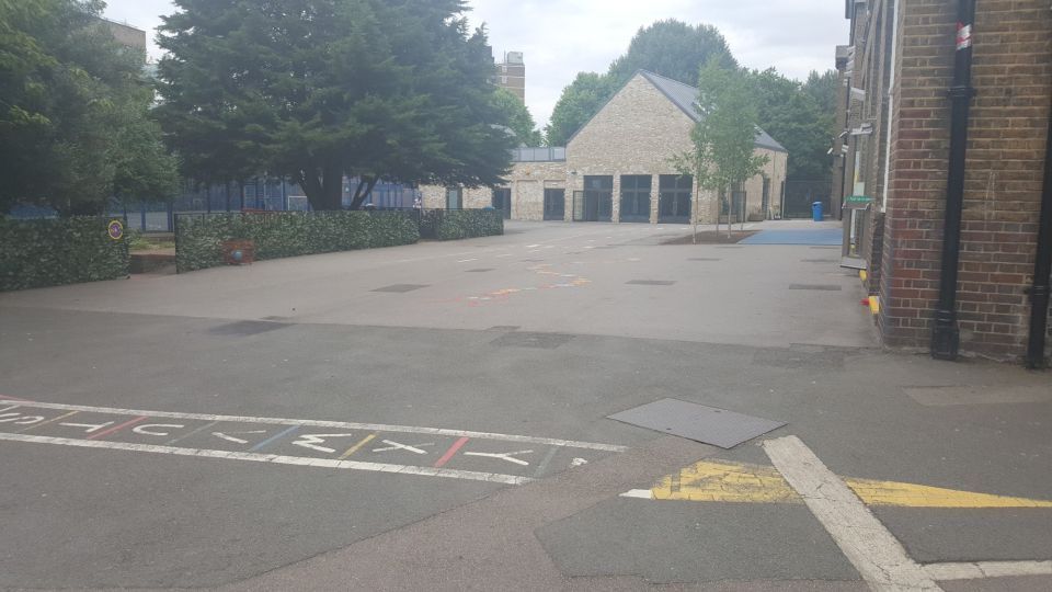 Schoolyard with asphalt surface, hopscotch, and brick buildings. Trees and sky in background.