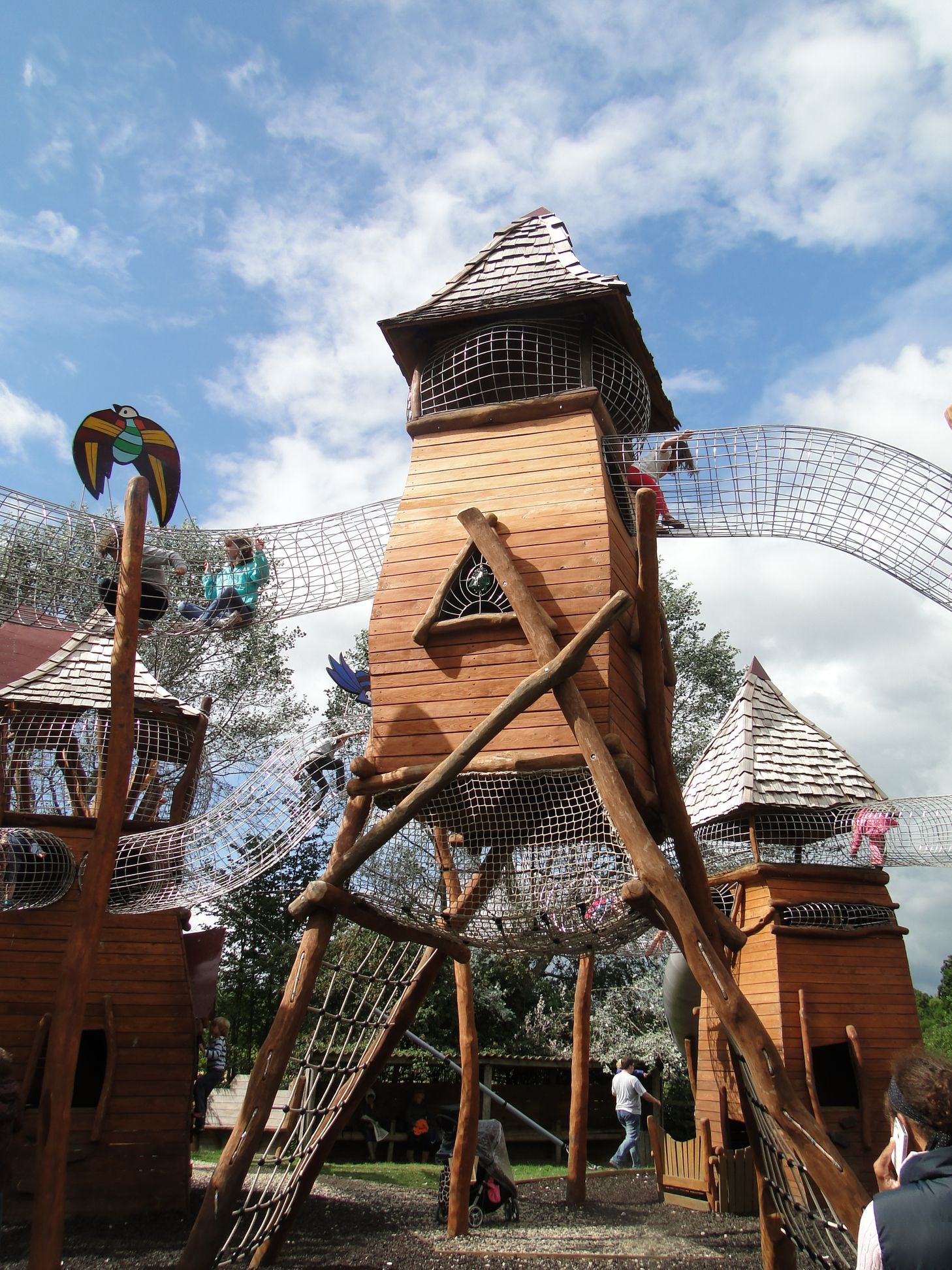 Playground with wooden towers, bridges, and children climbing on it under a blue sky.