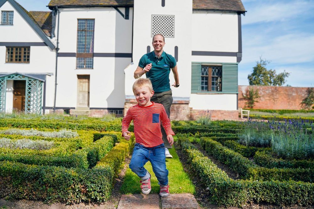 Father and son run through a garden maze in front of a white Tudor-style house.