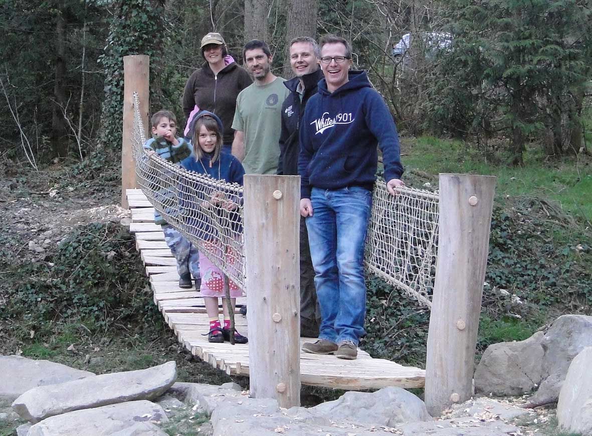 Group of people standing on a wooden bridge in a wooded area. Smiling, posing for the photo.