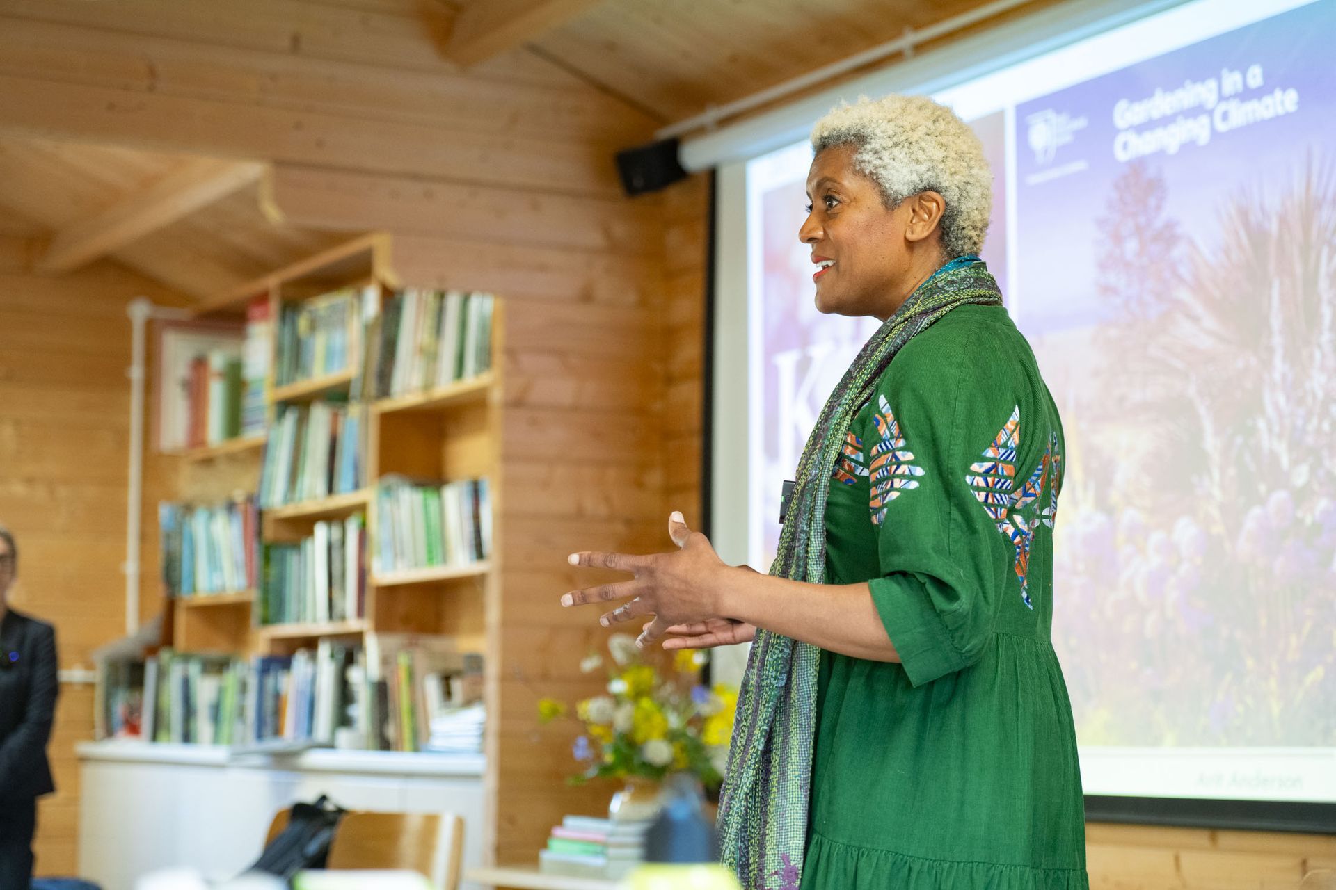 Woman with gray hair in green dress speaking in front of a projection screen.