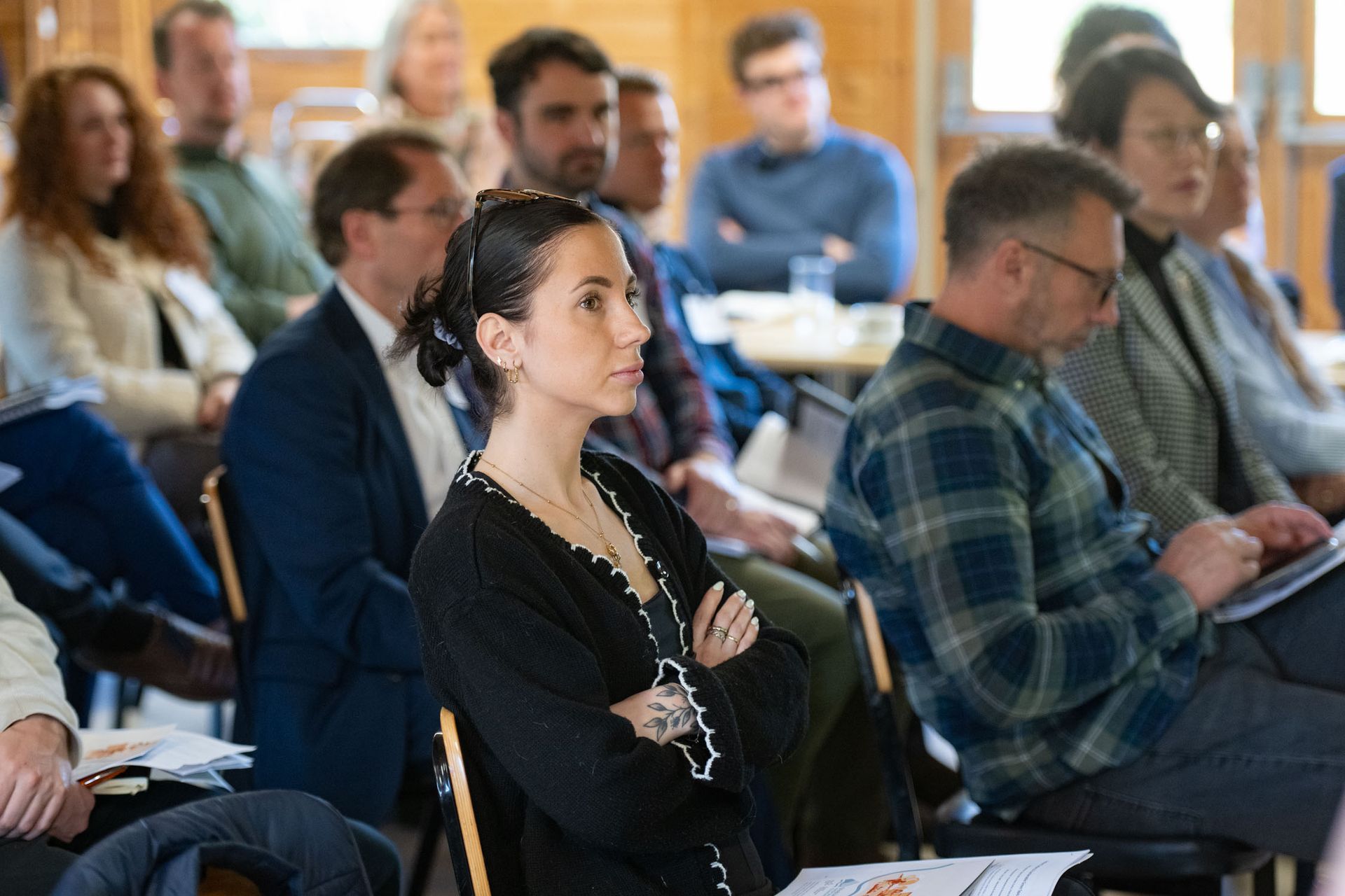 Audience in a meeting, focused on a woman with arms crossed, inside a wooden room.