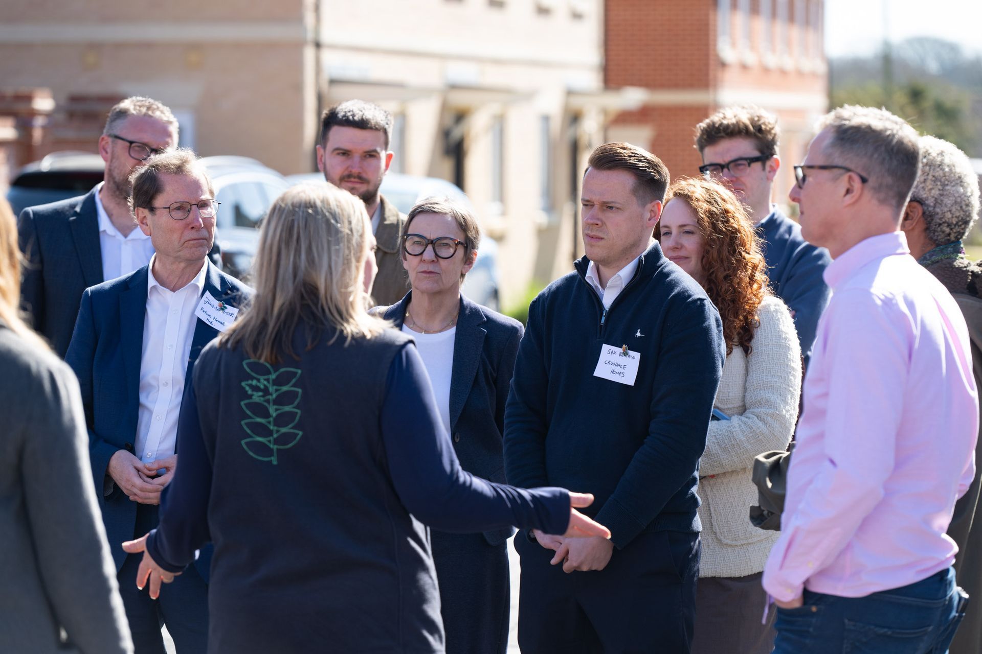A group of people listen to a woman gesturing outside buildings on a sunny day.