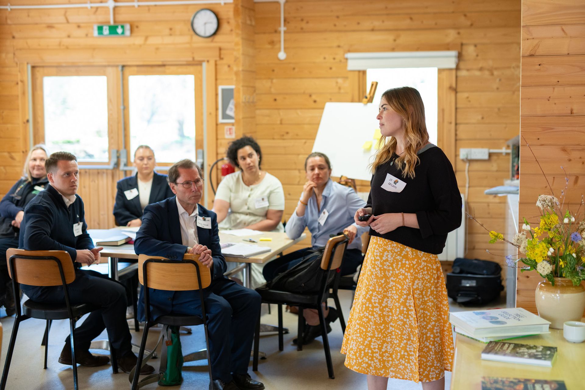 Woman presents to a group seated at tables in a wooden-paneled room.