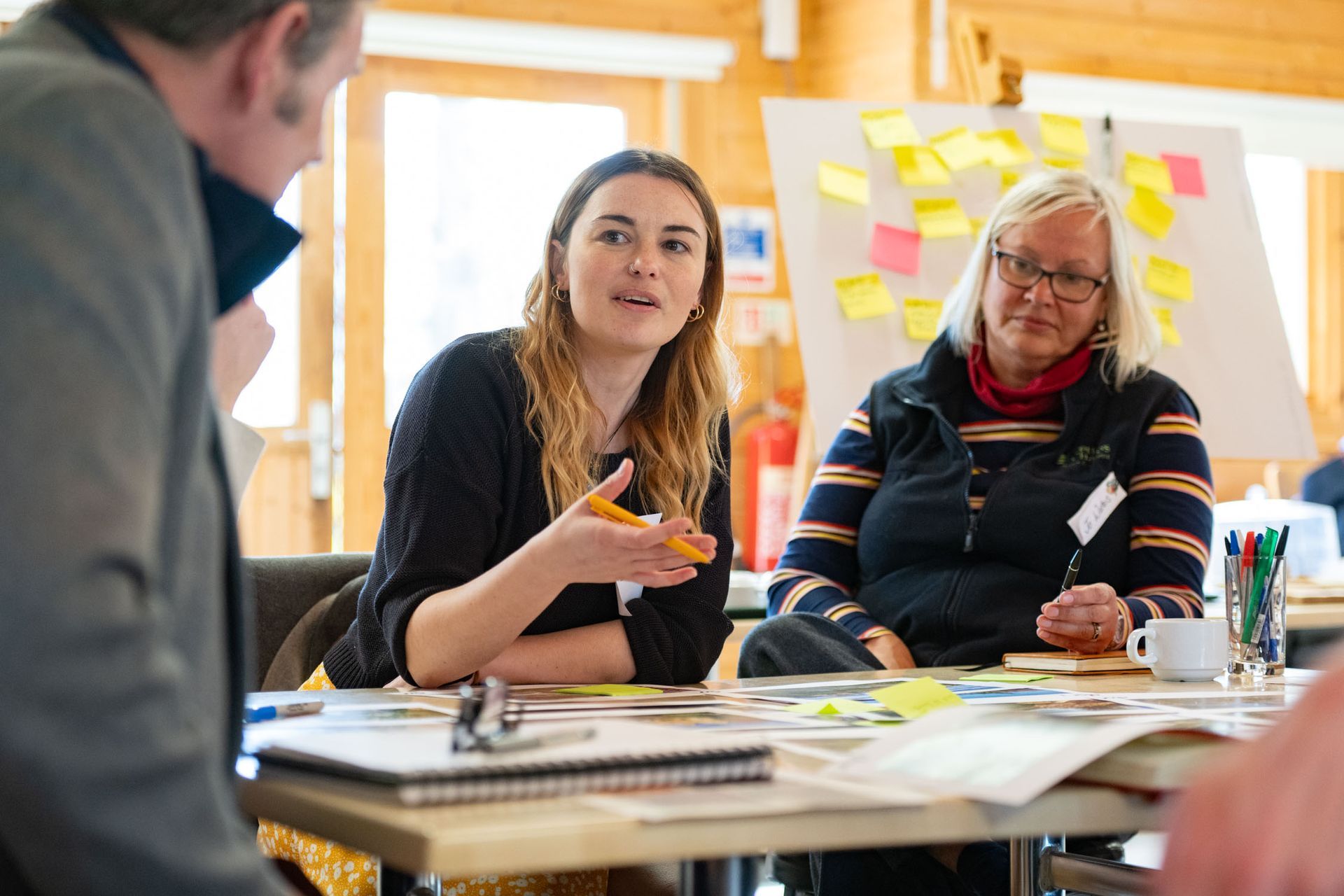 Three people at a table, one speaking with hand gestures, others listening in a brightly lit room; brainstorming session.