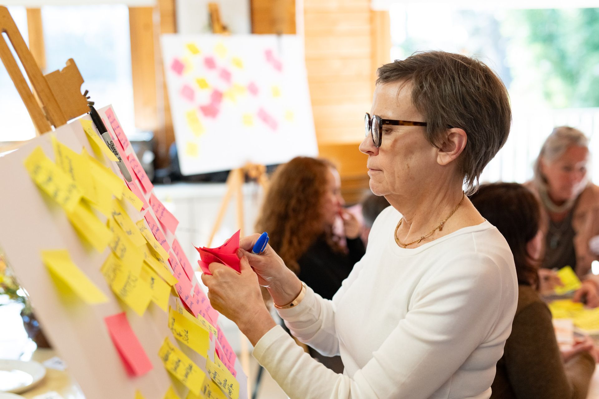 Woman with glasses placing sticky notes on a board during a workshop, others in the background.