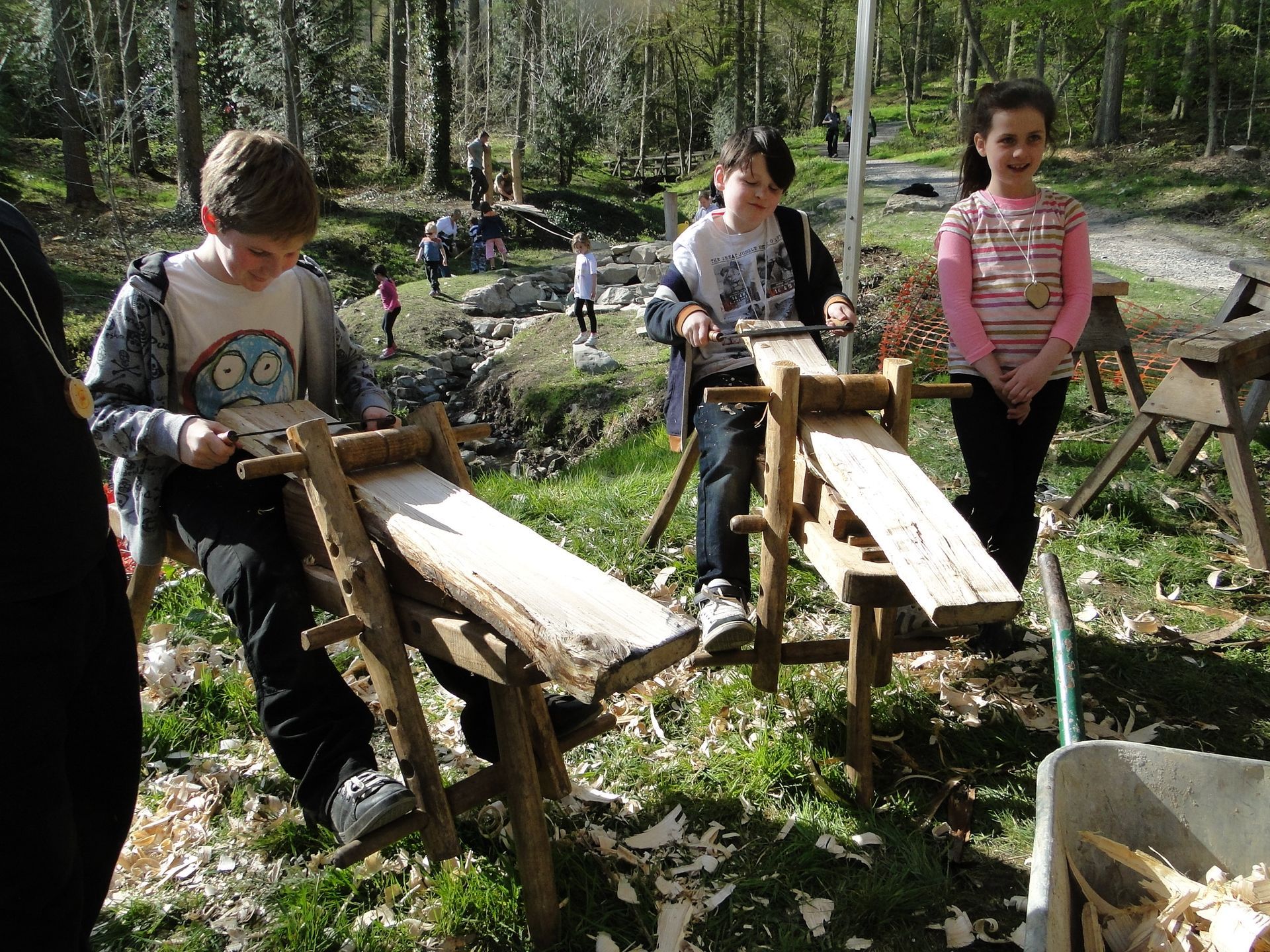 Children using wooden shave horses outdoors, shaving wood to build a bridge across a stream 