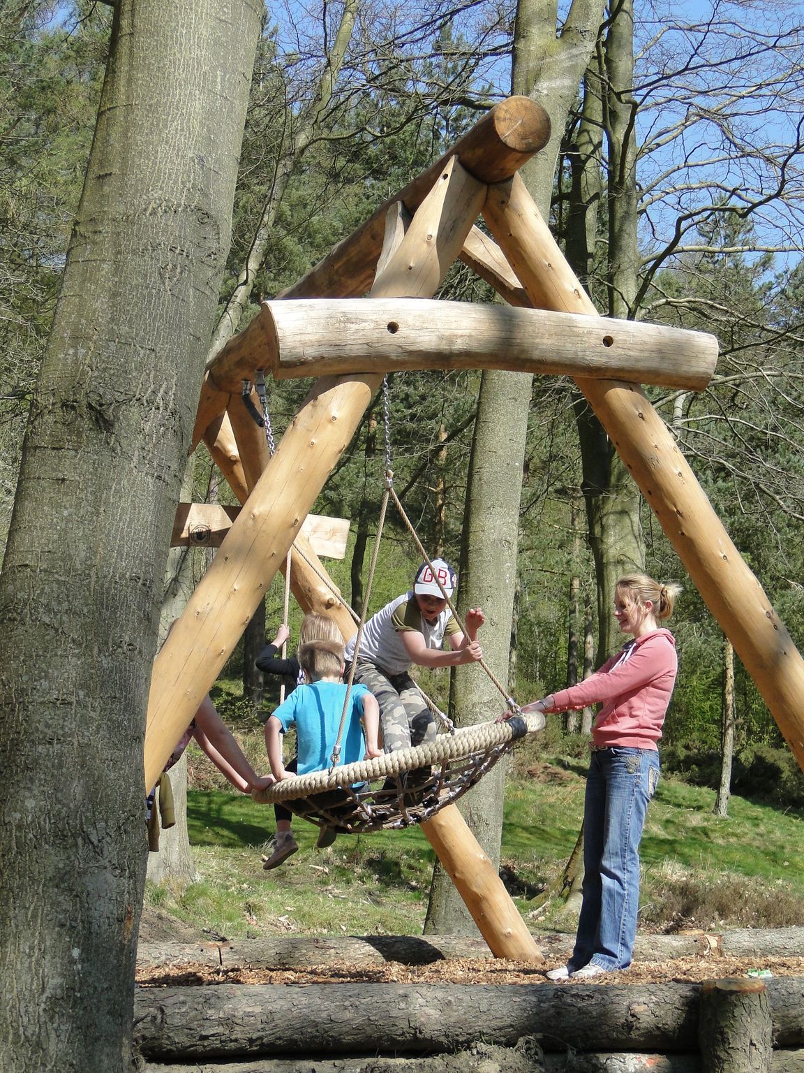 Children playing on a rope swing supported by a wooden A-frame structure in a forest.