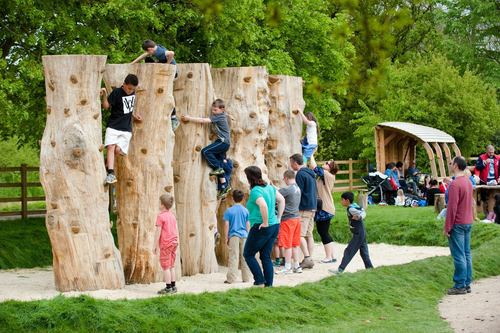 Children climb wooden structures at an outdoor playground; other kids watch and a few adults supervise.