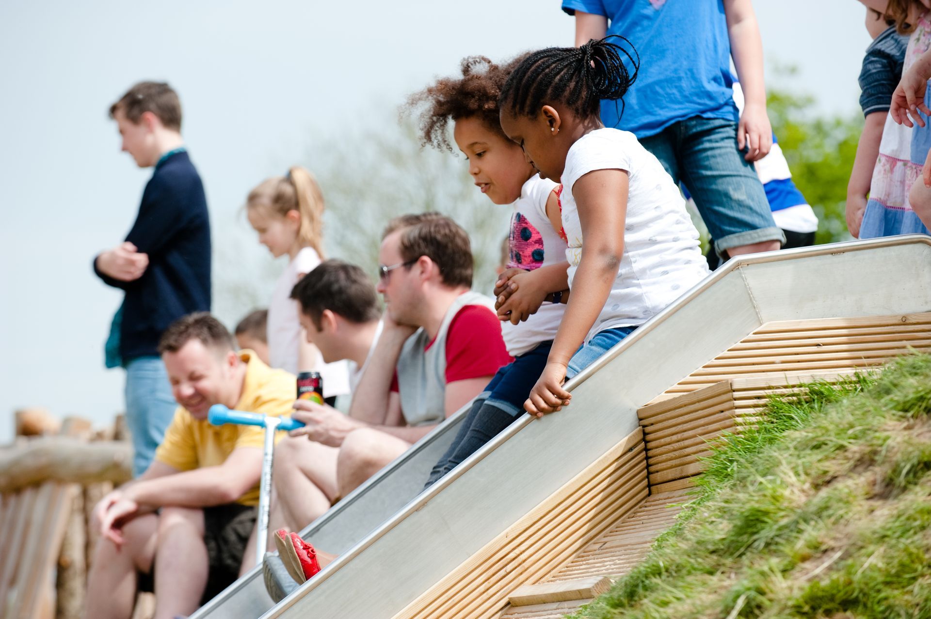 Children and adults watch at a playground. Two girls look down at a slide. Others sit nearby, some with scooters.