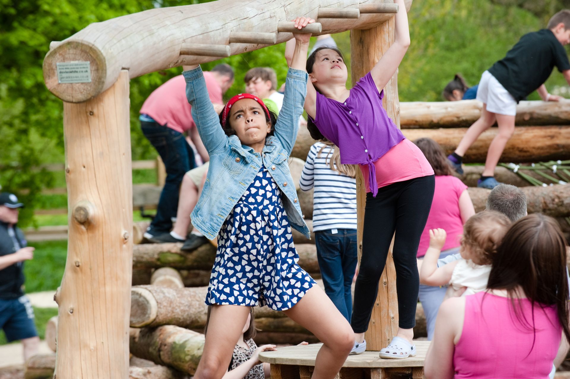 Children playing on a wooden playground structure. Girls in blue and purple tops climb and look up.