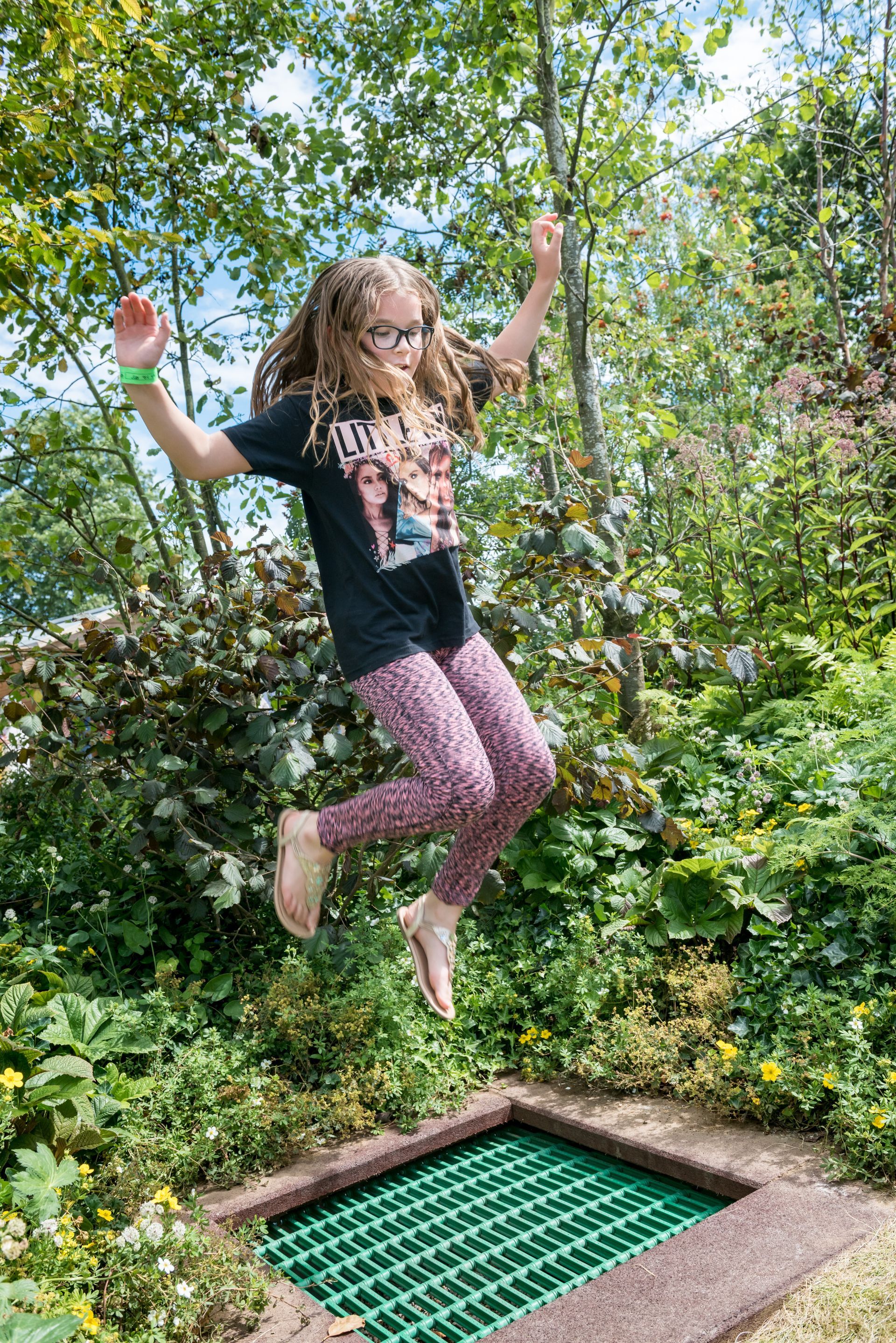 Young girl with curly hair jumps on a small trampoline outdoors, surrounded by greenery.