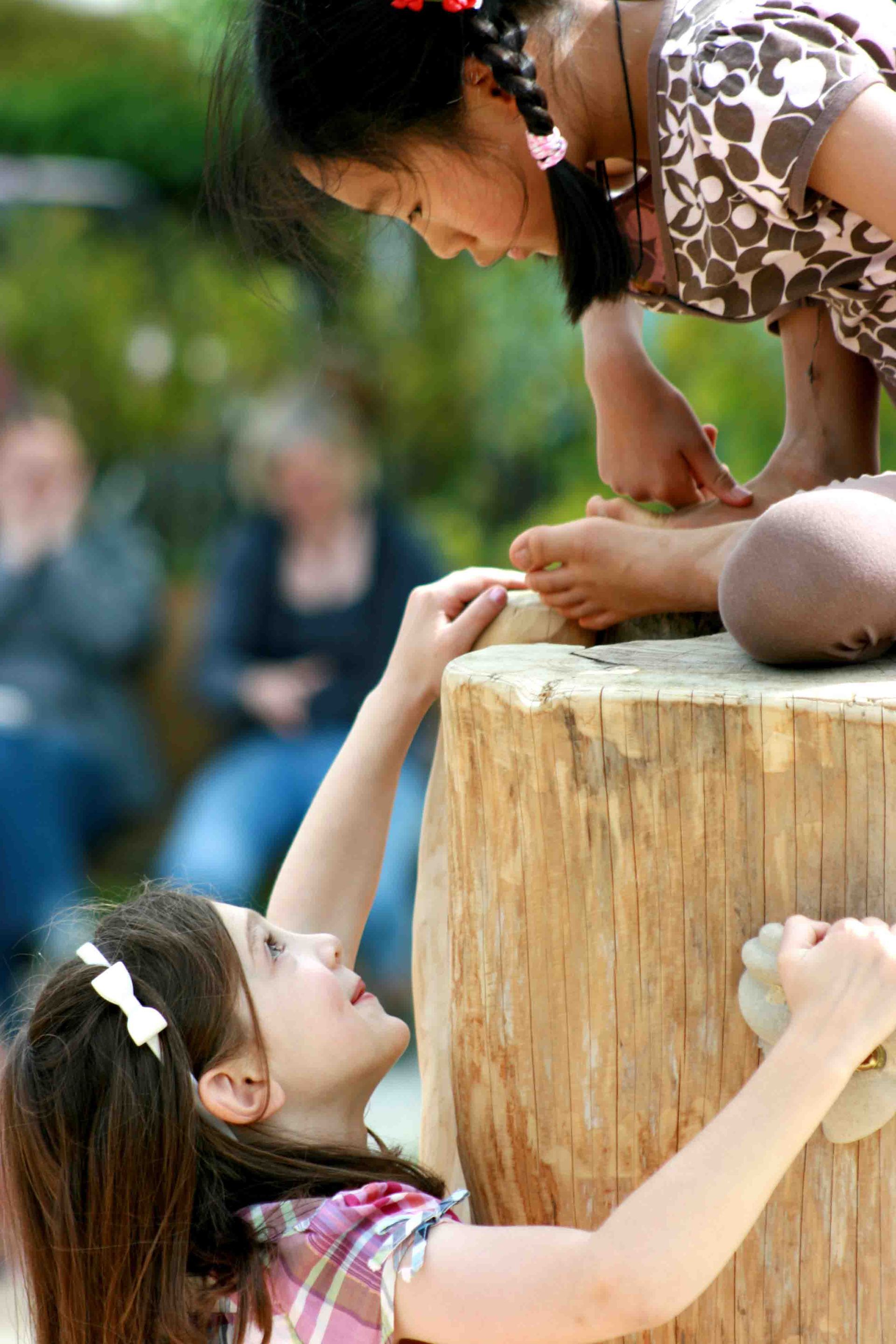 Two girls play outdoors, one helping the other climb a log; onlookers in background.