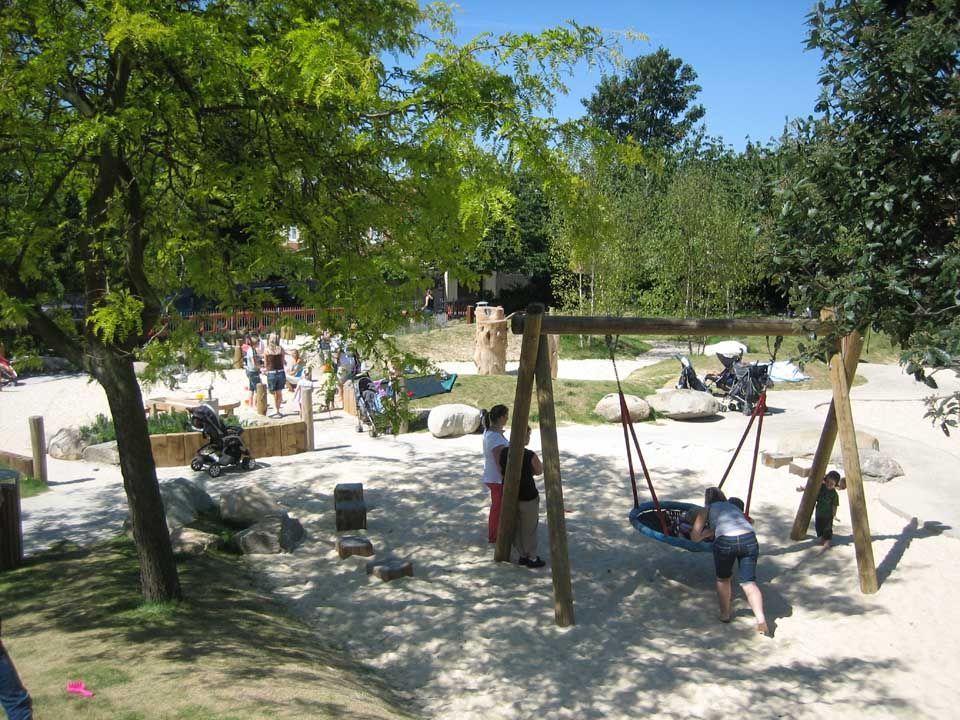 A sunny playground with swings, sand, trees, and children playing.