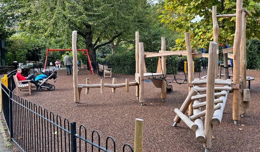 Playground with wooden climbing structures, swings, and people in the background.