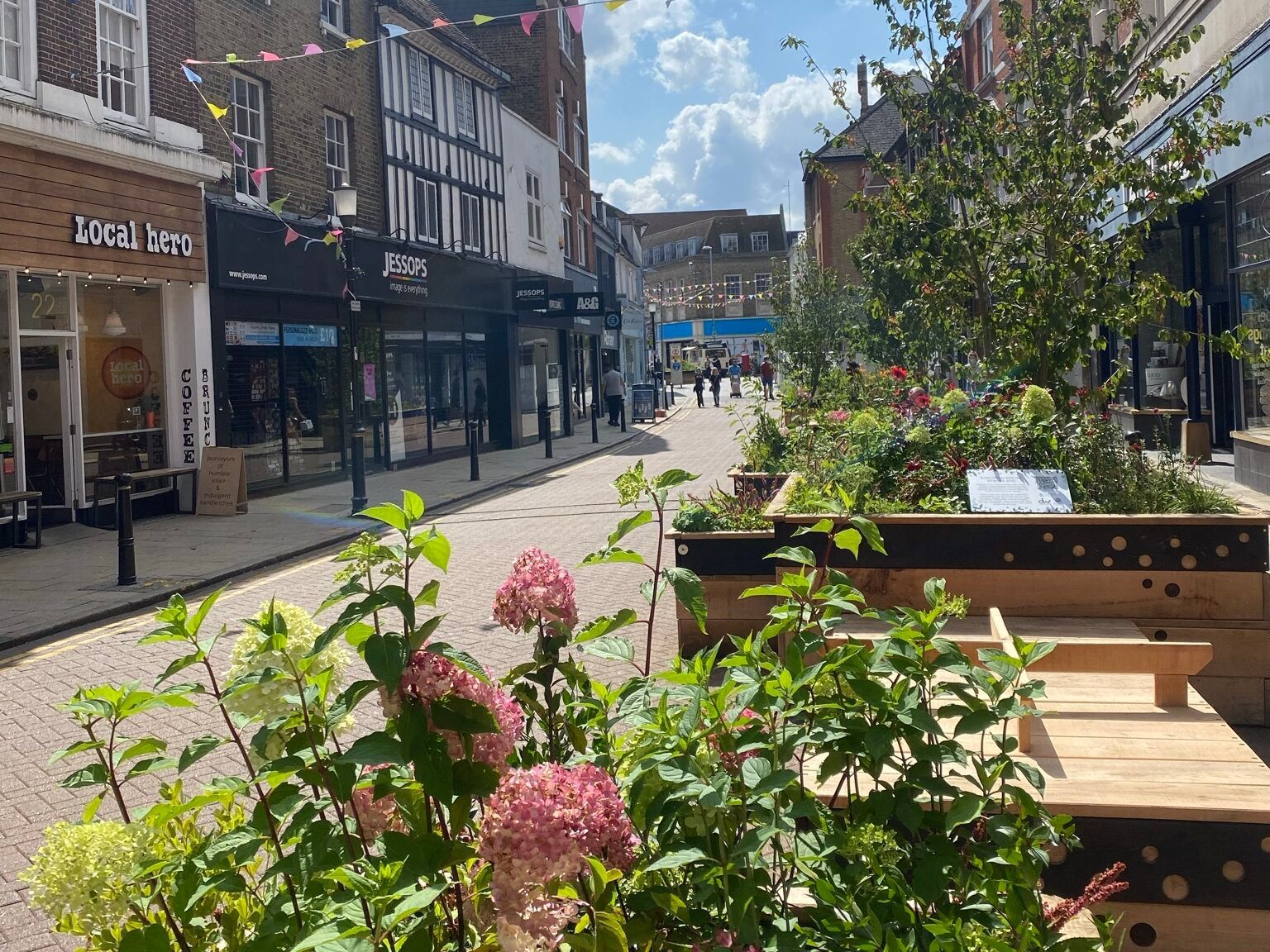 Street with flowers and plants. Shops line the street with people walking. Bright sunny day.