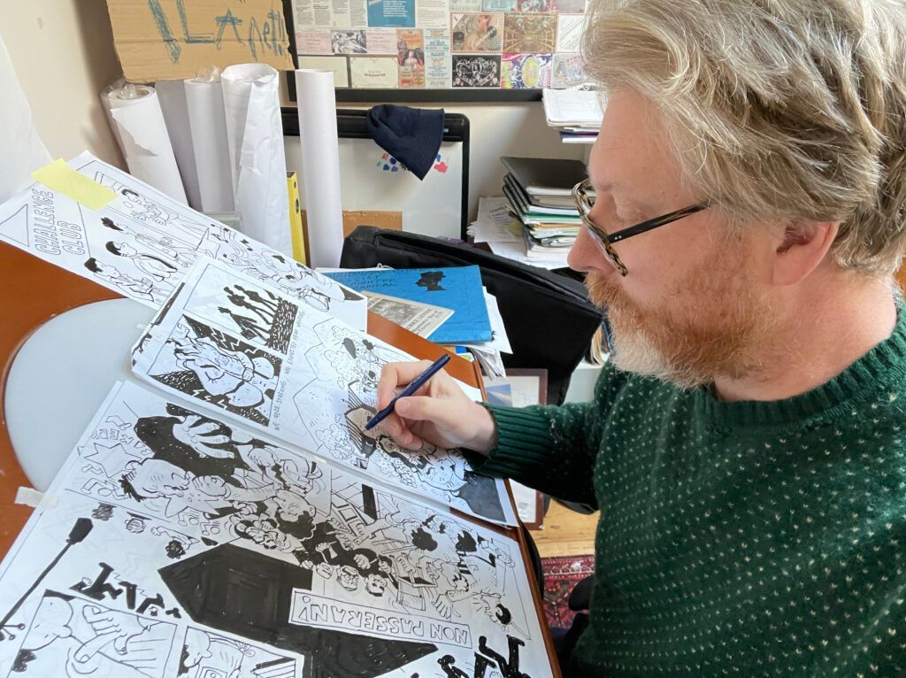 Man with glasses draws on a large black and white illustration, at a cluttered desk.