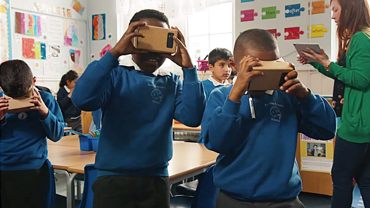 Children in a classroom wearing VR headsets, teacher holding tablet.