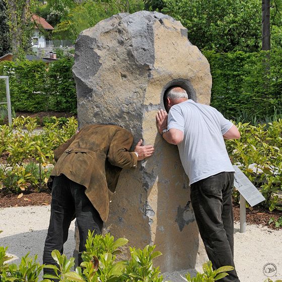 Two men looking through a large, stone sculpture with a hole; outdoor setting.