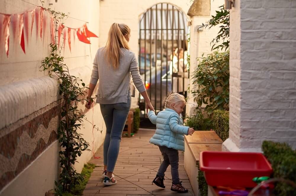 Woman and toddler walking down a brick alley, holding hands. Red bunting and a red bin are visible.