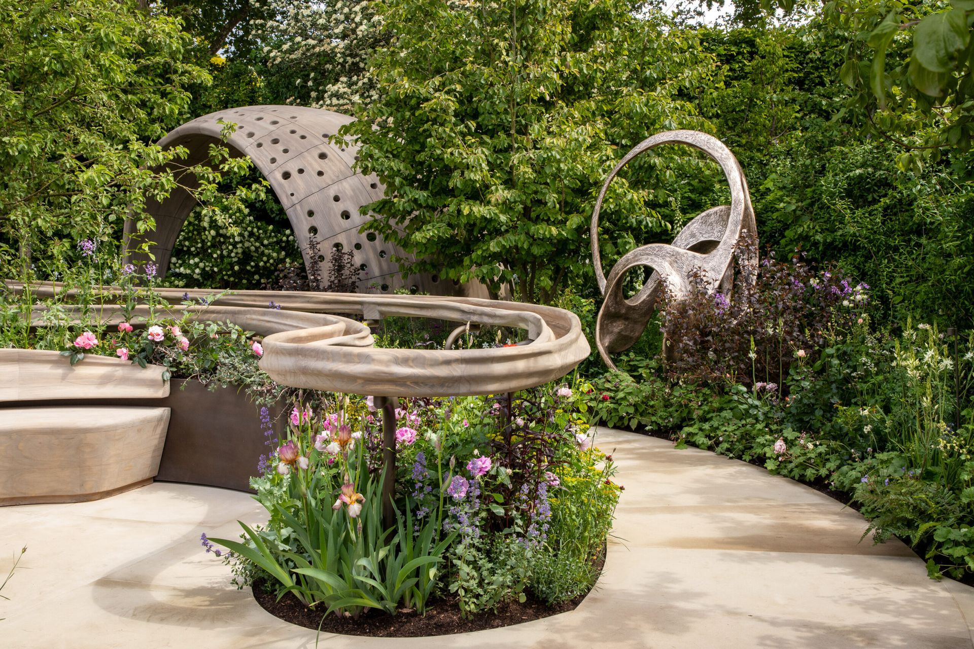 Arit Anderson - Modern garden with circular stone benches, a perforated metal sculpture, and lush greenery and flowers