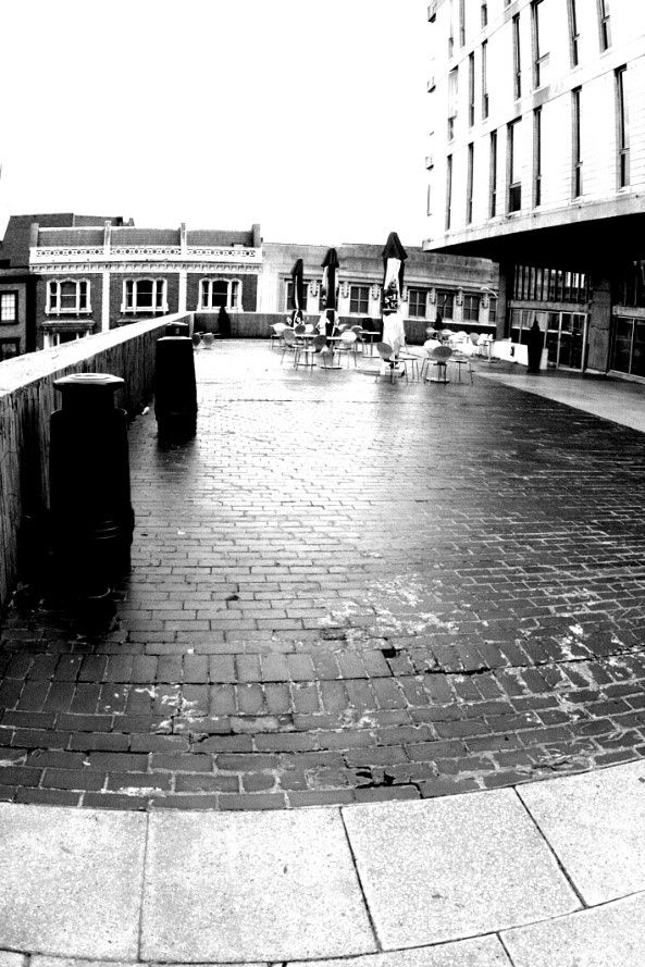 Brick patio with tables and umbrellas, next to a modern building. A street with older buildings is in the background.
