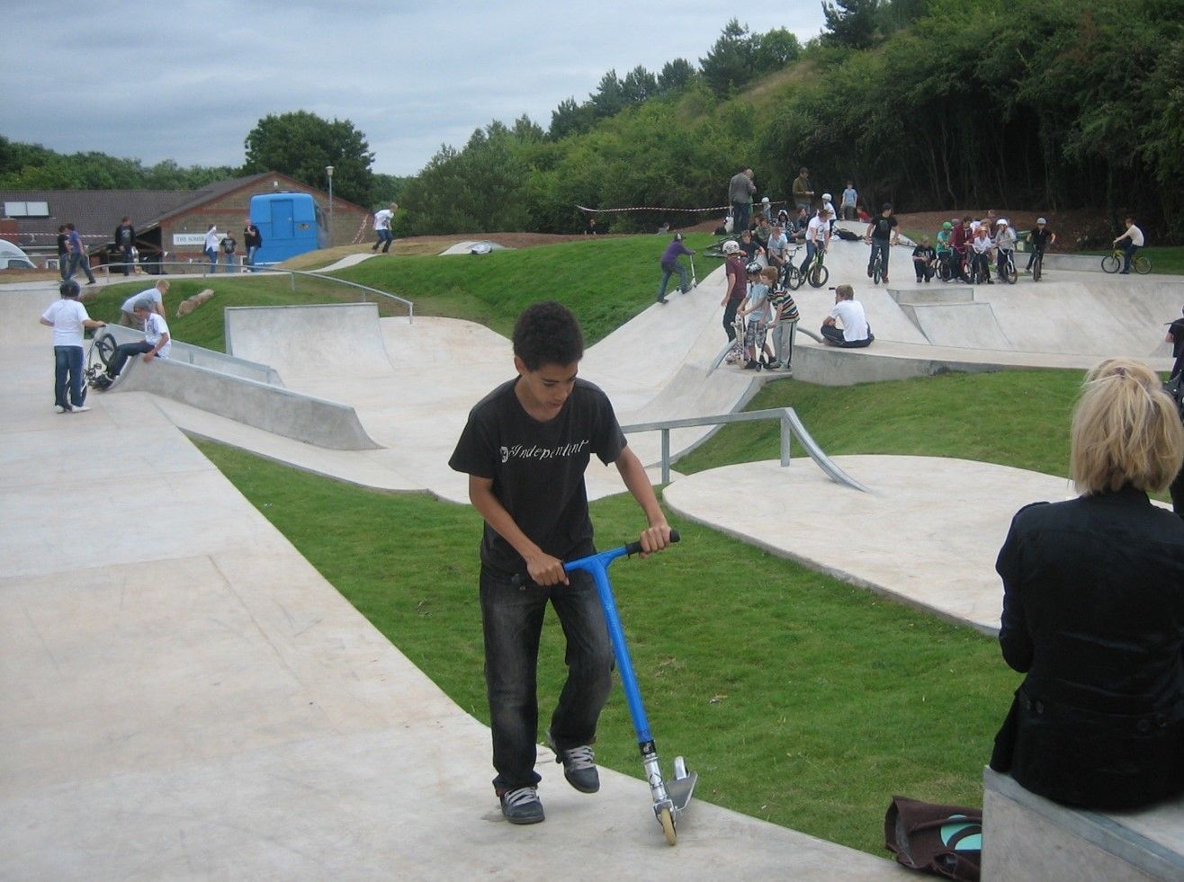 Boy on scooter at a skatepark. People on ramps and grassy areas. Concrete and a building in the background.