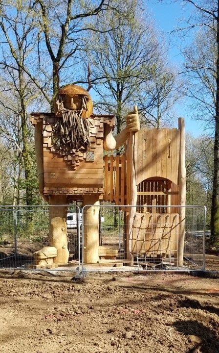 Wooden playground structure with climbing features, trees, and a fence.