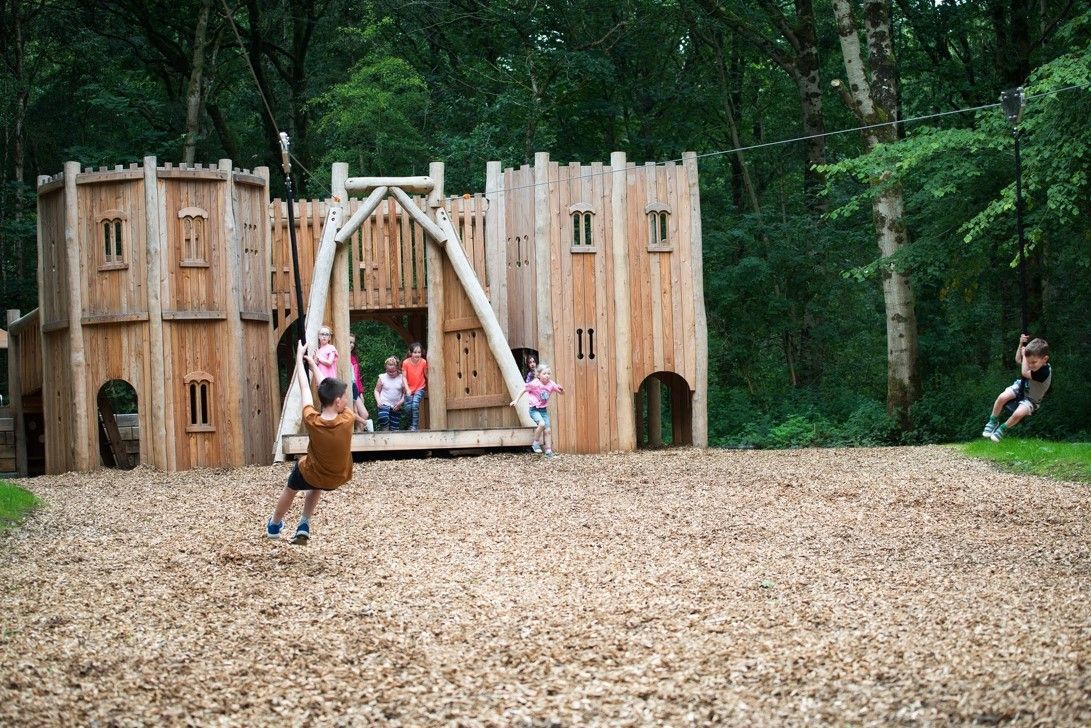 Children on zip lines in a wooden castle playset, set in a forest.