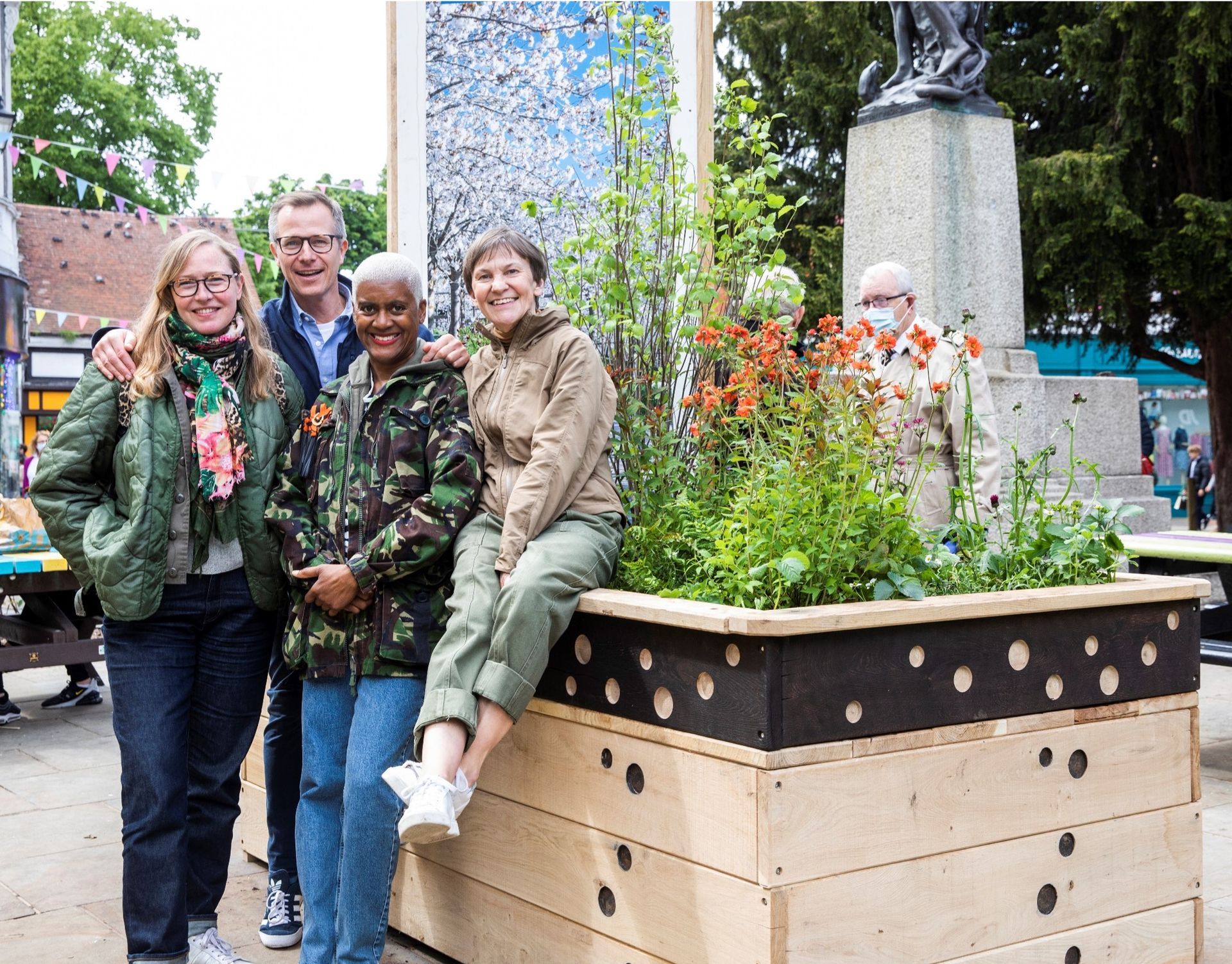 Group of six people posing next to a raised garden bed in a plaza with a statue.