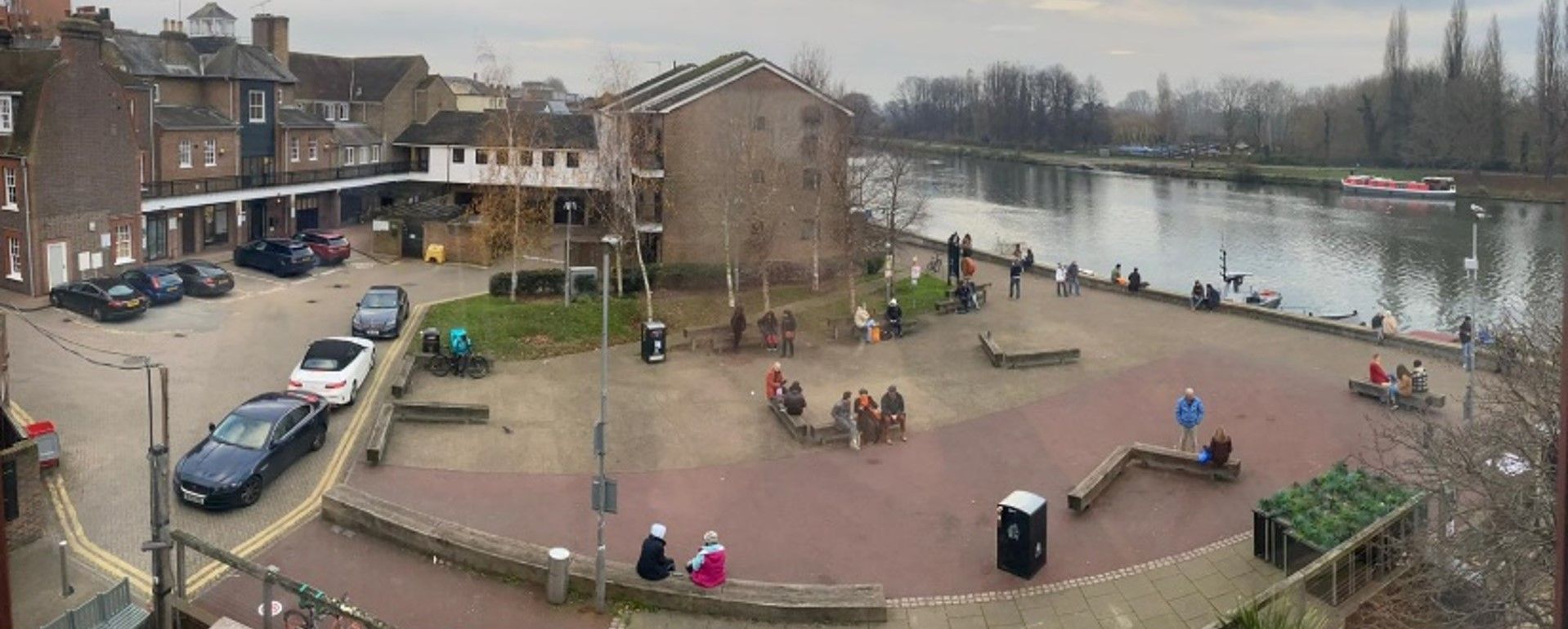A riverside scene featuring brick buildings, parked cars, and people by the water. Cloudy sky.