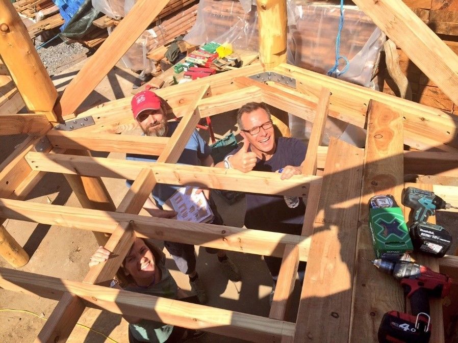 Three people smiling and giving thumbs-up, building a wooden structure outdoors, surrounded by tools and wood.