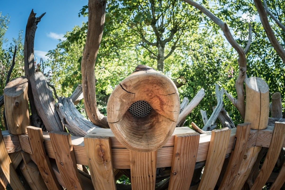 Wooden sculpture with a large horn shape, set against a fence and trees.