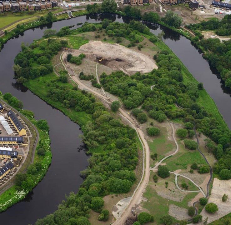 Aerial view of a green, tree-covered island in a dark river, with walking paths and a building in the background.