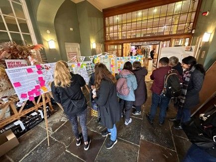 People gathered at a display with notes, indoors. Some are reading the notes, and others are looking on.