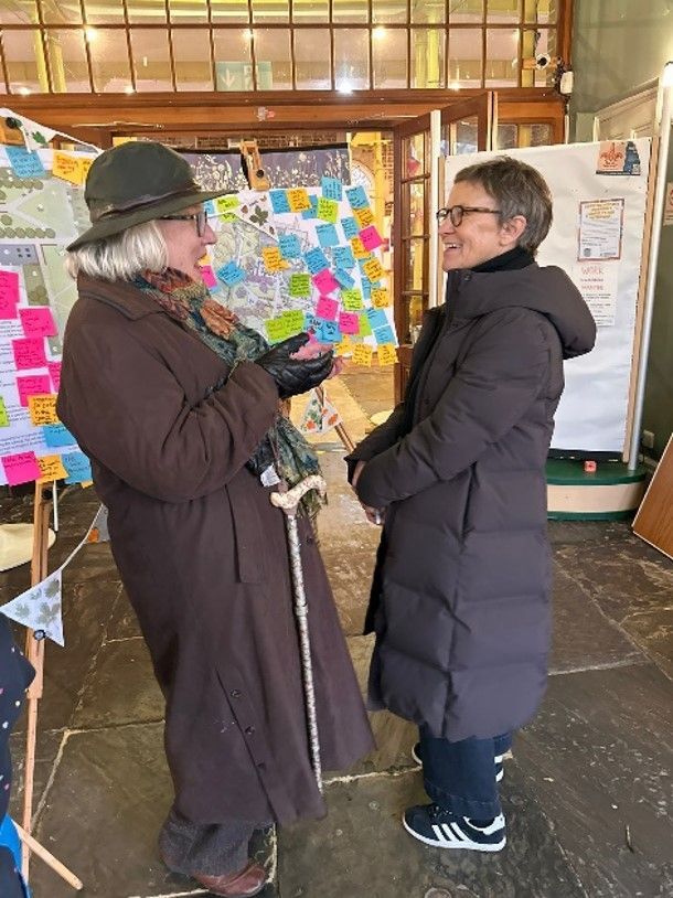 Two women talking in front of a display with colorful sticky notes. One wears a brown coat and hat.