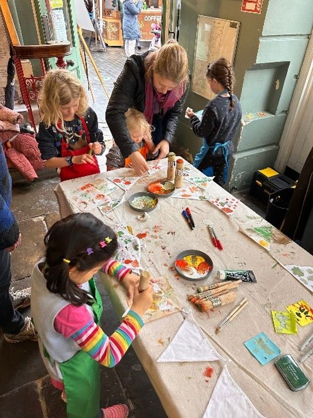 Children and an adult painting at a table. Colorful paint, flags, and supplies are visible.