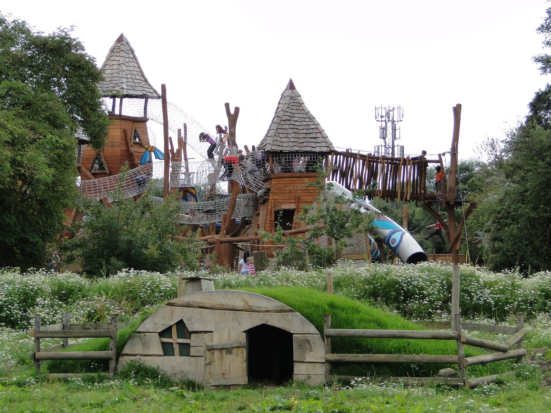 Playground with wooden structures, including towers and a small hobbit-like house with a grassy roof.