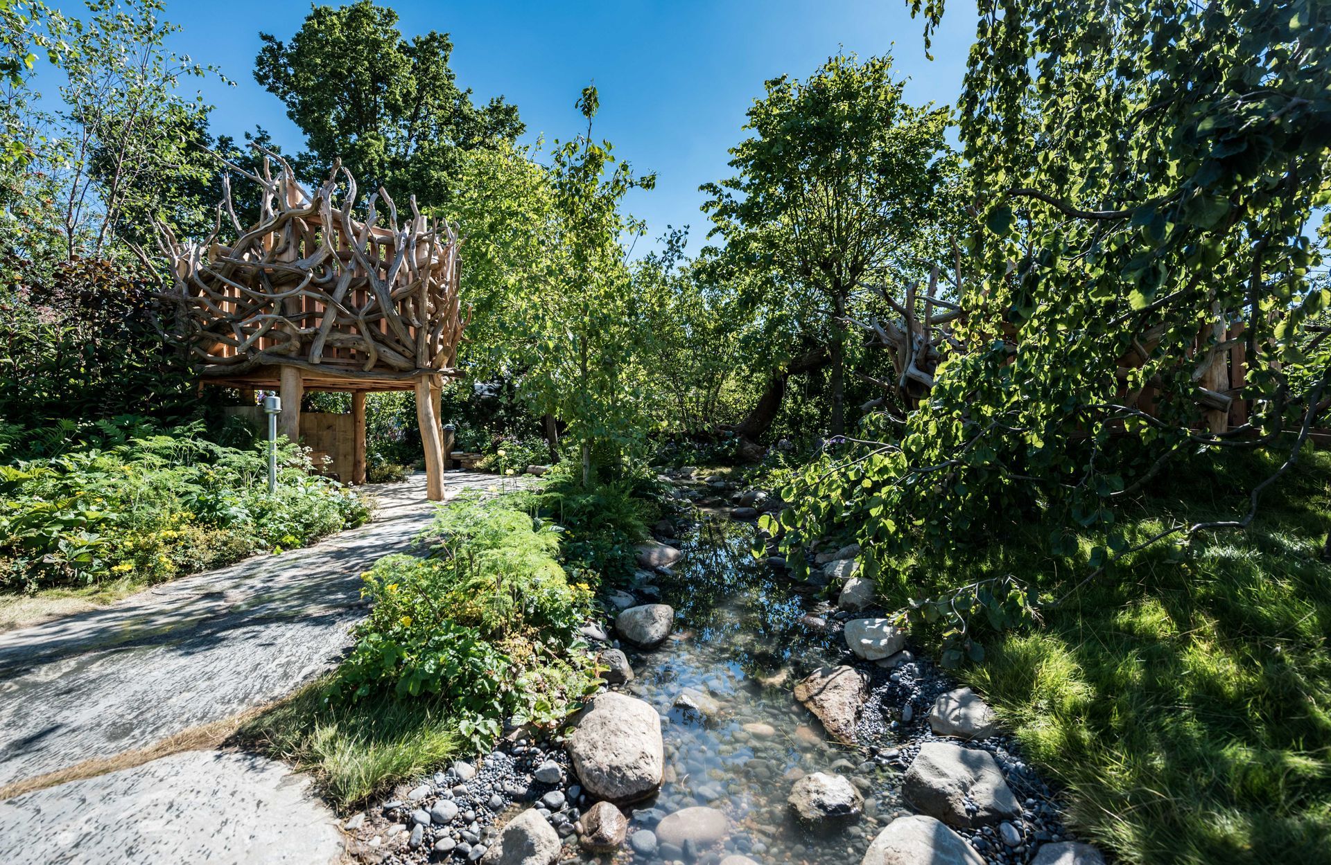 A wooden structure sits among lush greenery next to a rocky stream. Bright sunlight shines.