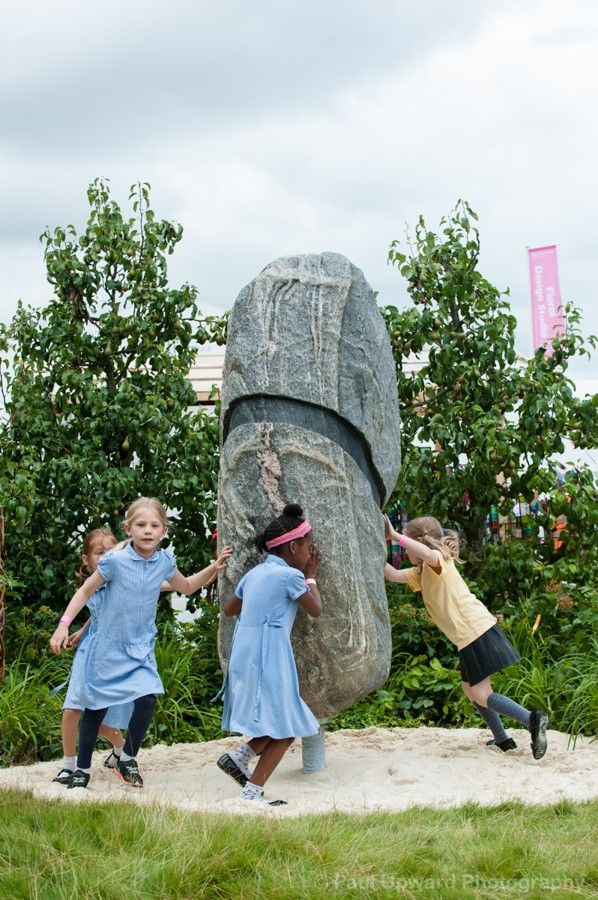 Children in school uniforms play near a large stone sculpture in an outdoor setting.