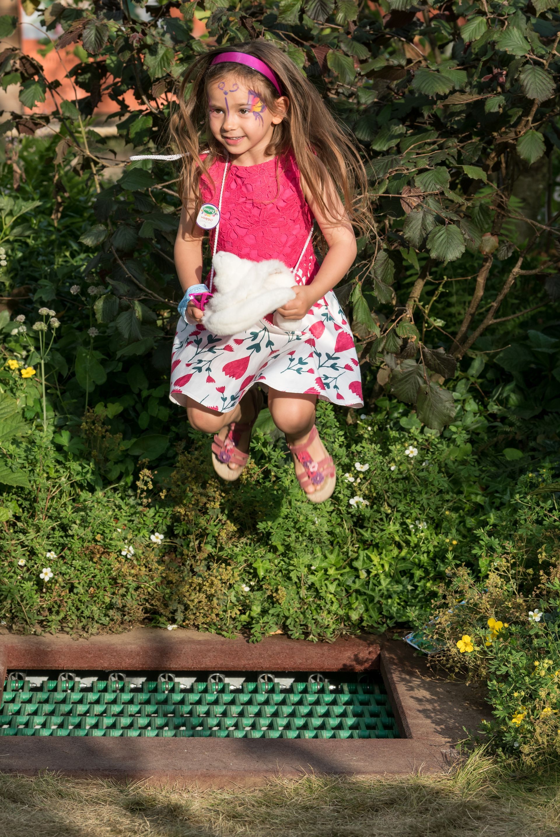 Girl jumps in the air, smiling, in a garden. She wears a pink top, floral skirt, and headband.