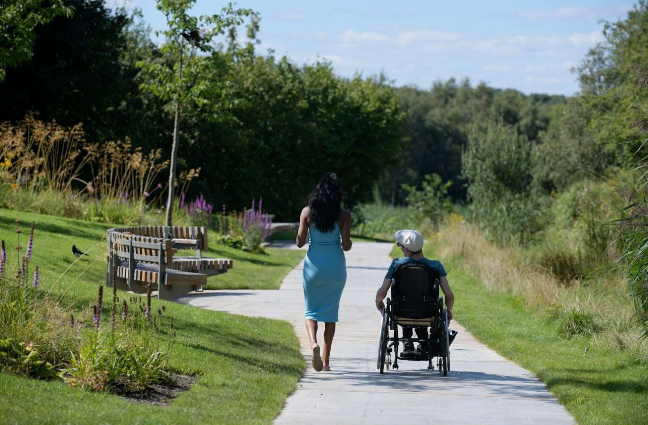 Woman in blue dress walks with a person in a wheelchair on a park path, sunny day.
