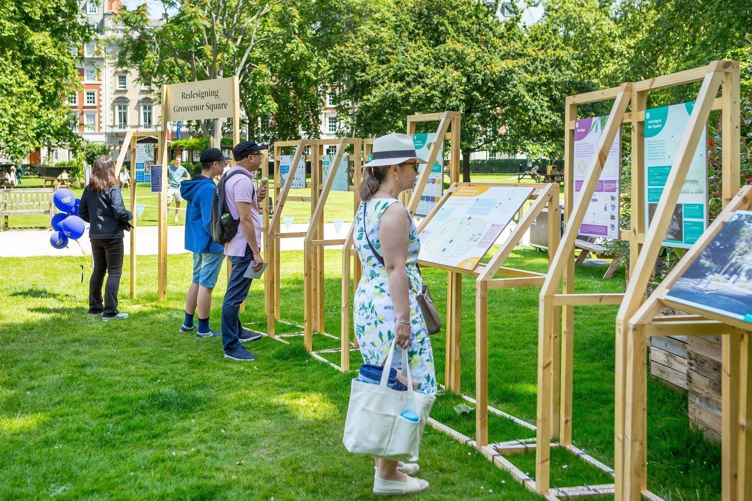 People viewing informational displays in a park on a sunny day. Wooden frames hold signs on green grass.