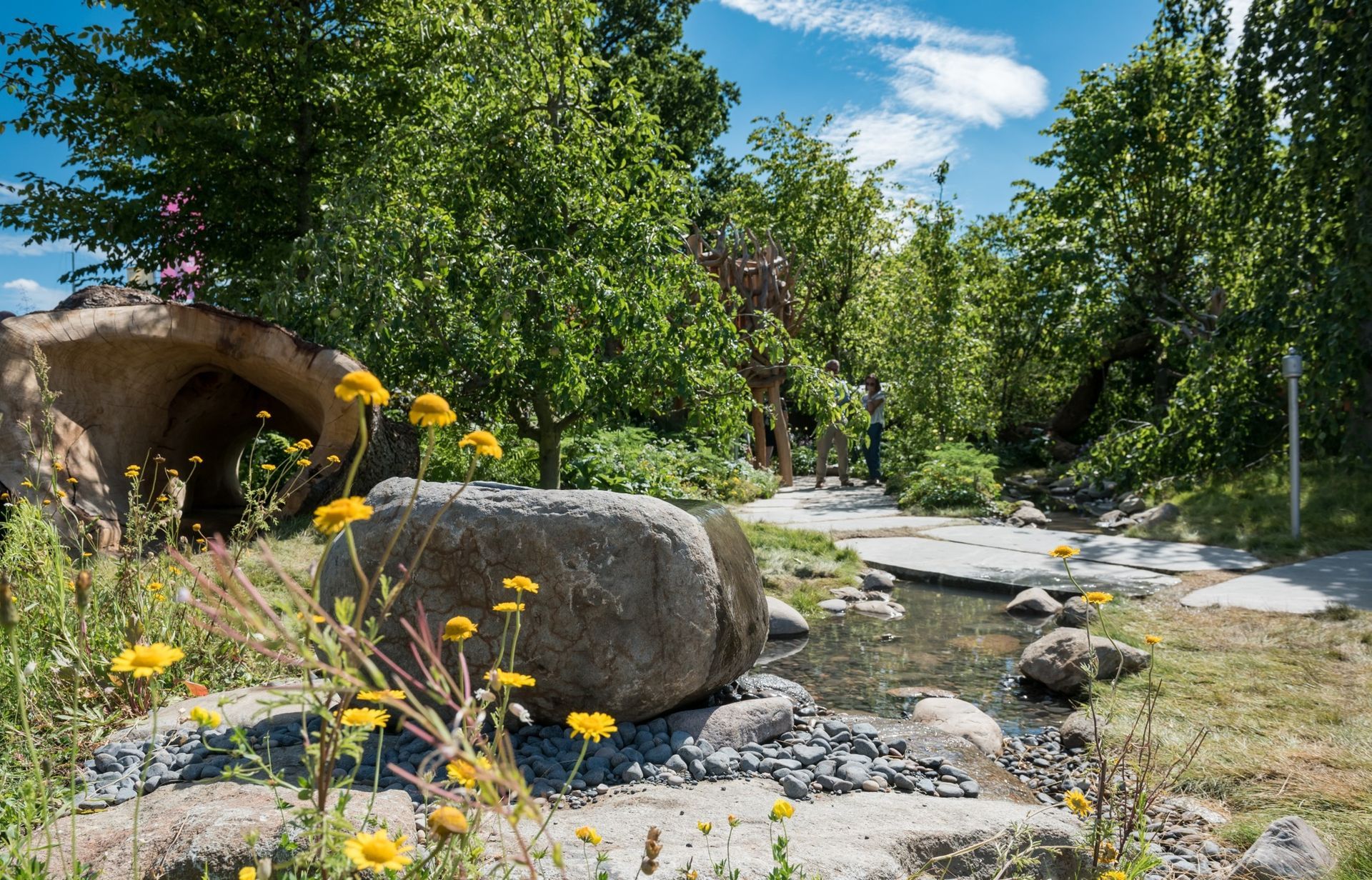 A rock-lined stream with yellow wildflowers, a concrete tunnel, and lush green trees under a blue sky.
