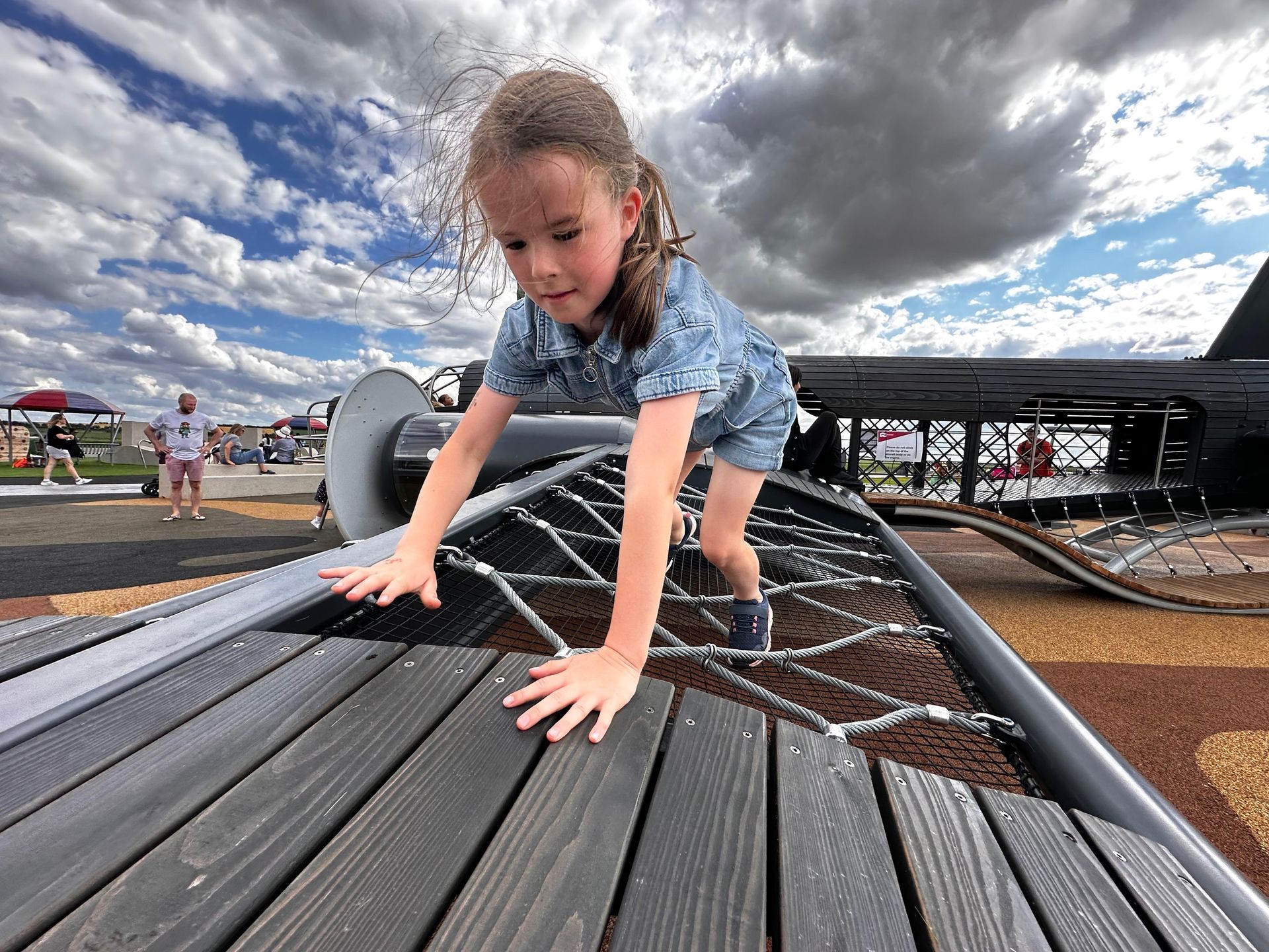 Girl climbing on a playground structure with a net, outdoors under a cloudy sky.