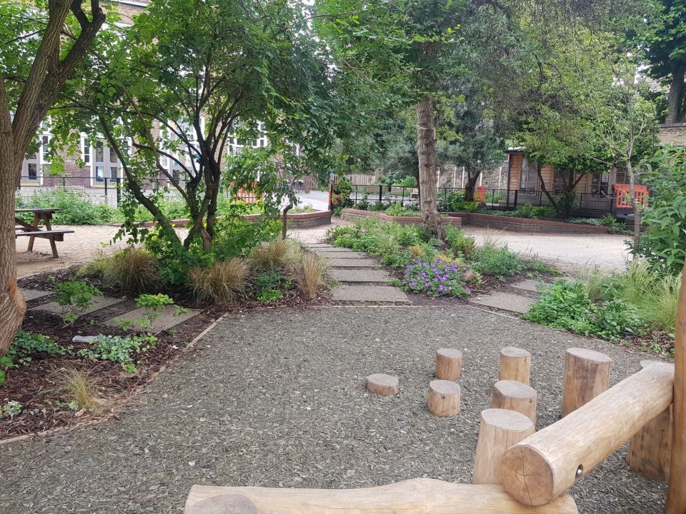 A child's garden with a gravel area, stepping stones, and wooden structures surrounded by plants and trees.