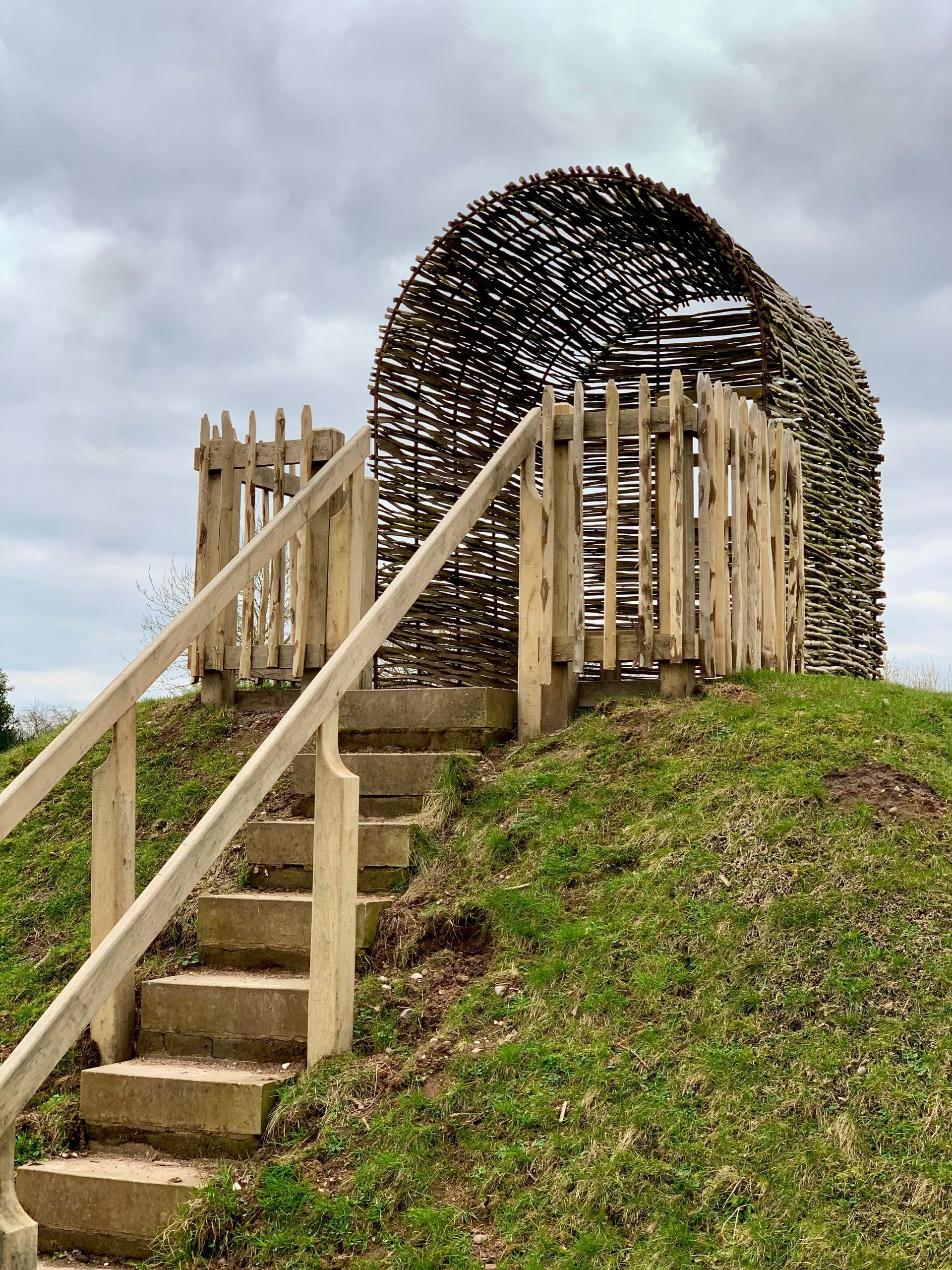 Wooden stairs leading up to a woven structure on a grassy hill under an overcast sky.