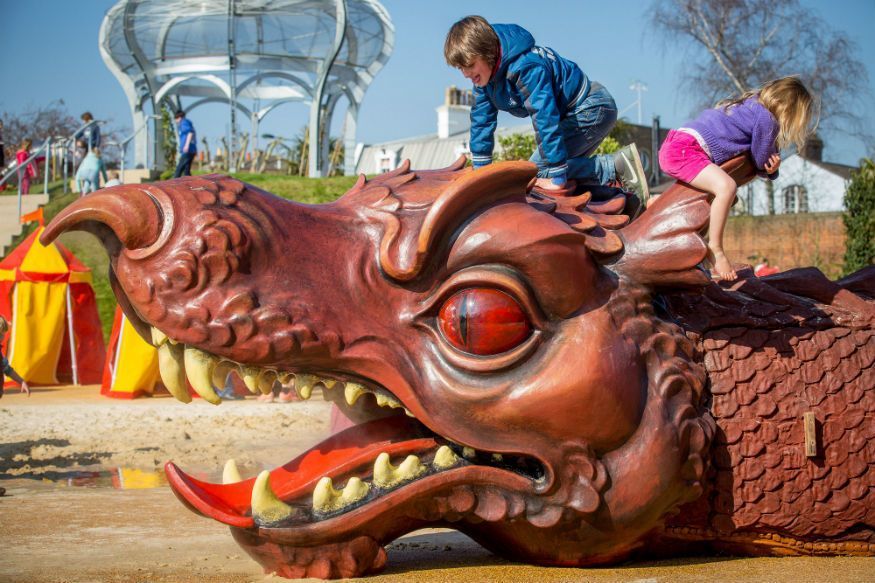 Children playing on a large, red dragon sculpture with open mouth, outdoors in a park.