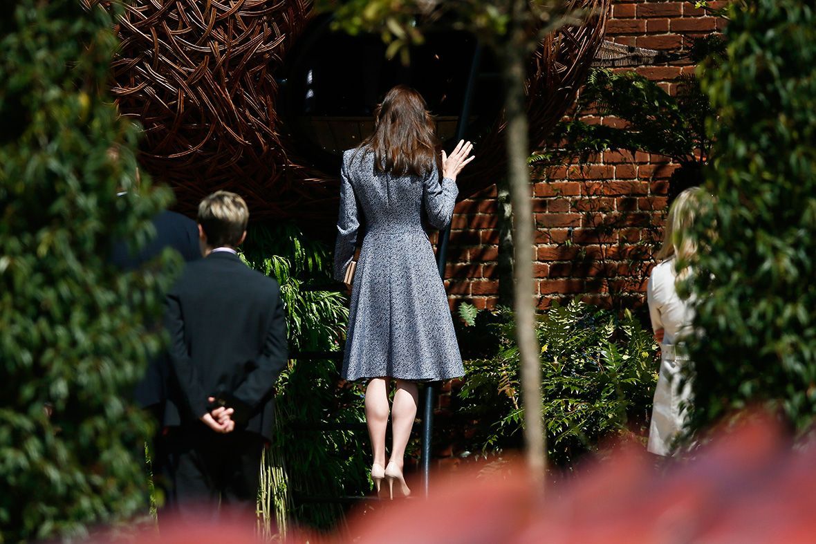 Woman in a blue dress waves, climbing steps to a brick building; two people stand nearby, with greenery framing the scene.